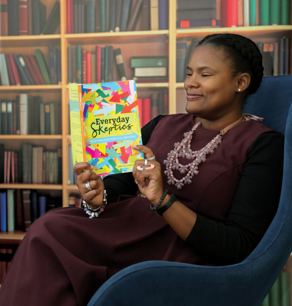 Alydia Smith, the editor of 'Everyday Skeptics', sits in a chair looking at the text of the book, holding it so that the cover is clearly visible. It features multicoloured arrows going in many directions.