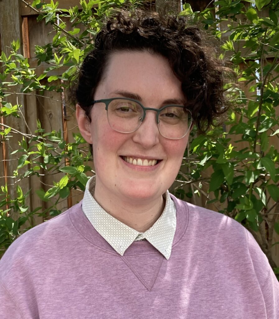 A photo of Bailey, a white non-binary person with short dark curly hair and glasses.